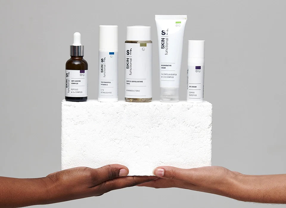 Collection of skincare products on a white block with two hands reaching towards them against a gray background.
