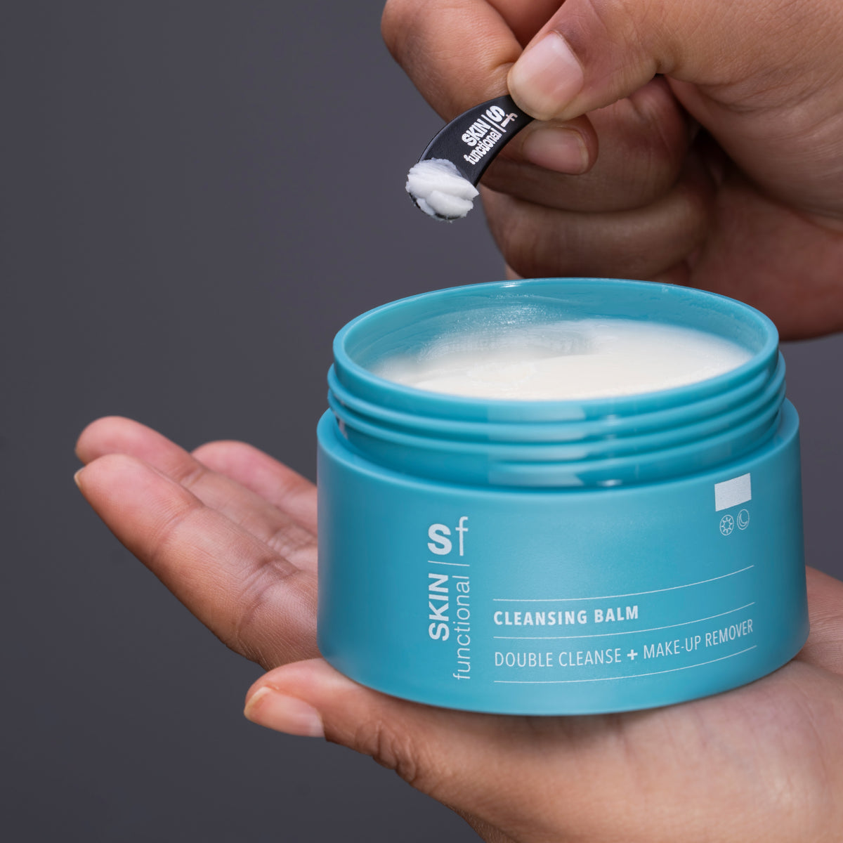 Blue jar of cleansing balm with a hand holding a applicator against a gray background